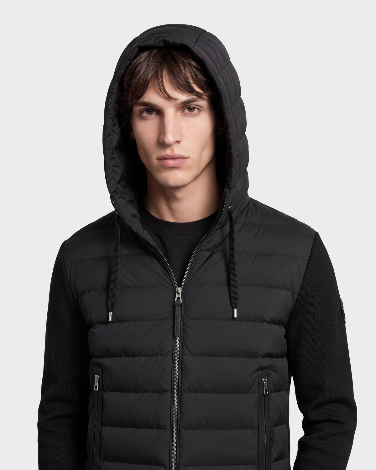 A young man stands facing forward against a plain light background, sporting the Rudsak RIVER HV - H125503 Black hybrid hooded down jacket filled with RDS-certified down, embodying a modern interpretation of Harvard THE COOP style.
