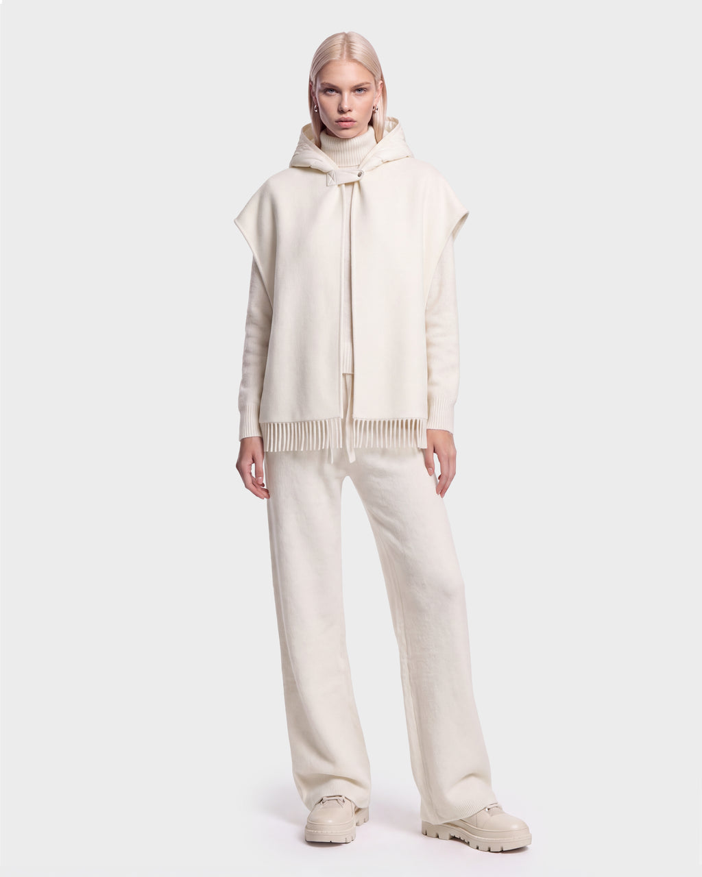 A person with straight blonde hair wears the ROXANE - 8526677 Vanilla poncho by Rudsak—an all-cream hooded fringe poncho styled with a long-sleeve top, wide-leg pants, and chunky shoes, standing against a plain light background.