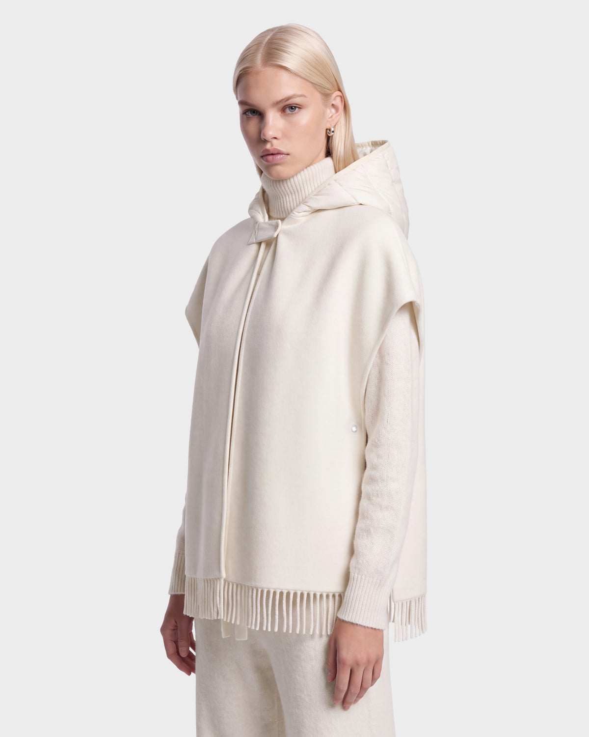 Someone with long blonde hair showcases the ROXANE - 8526677 Vanilla hooded poncho by Rudsak, featuring fringe detailing in a cream shade. The poncho is layered over a high-neck sweater and matching pants, all set against a plain light background.