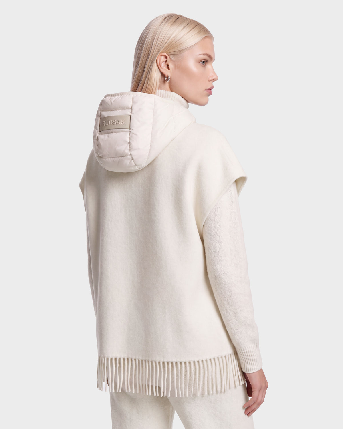 A woman with straight blonde hair wears the ROXANE - 8526677 Vanilla cream hooded poncho from Rudsak, featuring fringe detail at the hem and matching pants. A small label accents the hood as she faces away, gazing left against a light gray background.