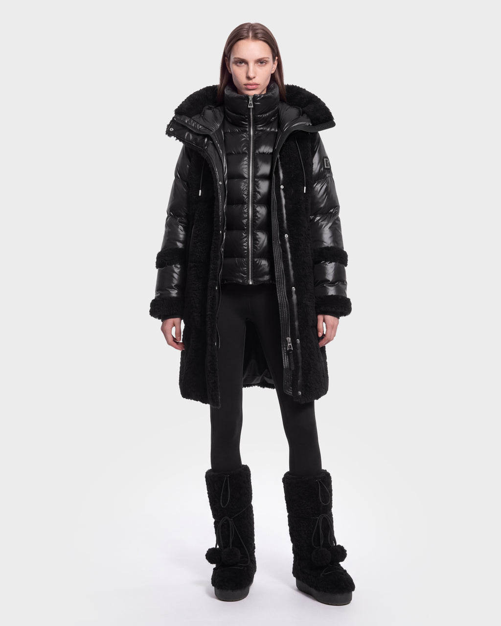 A person stands facing forward, dressed in the Rudsak SHELLEY - 8126729 Black long coat layered over a shiny black puffer jacket, paired with black leggings and tall black winter boots with pom-poms. The background is plain and light-colored.
