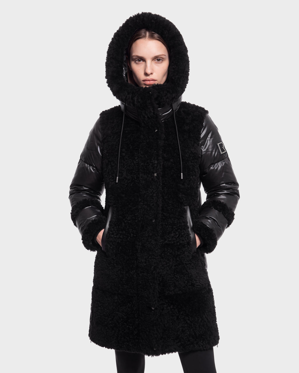 A person wearing the Rudsak SHELLEY - 8126729 Black coat, featuring shiny sleeves and a hood, stands with hands in pockets against a plain light background.