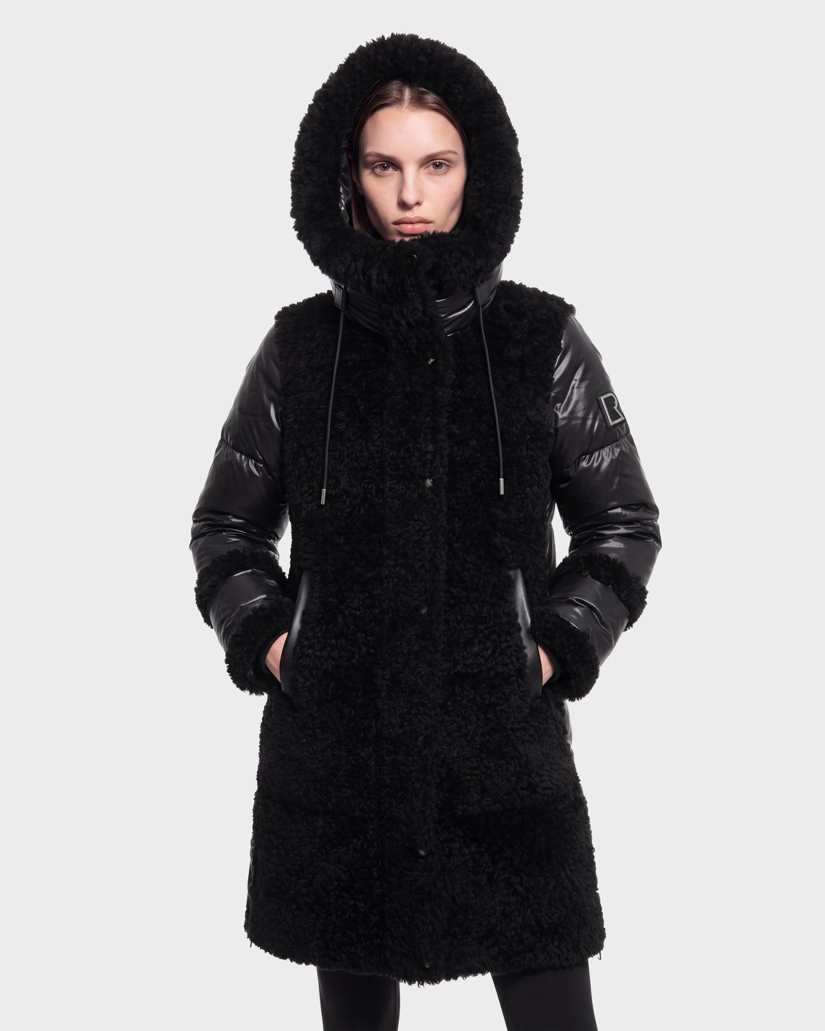 A person wearing the Rudsak SHELLEY - 8126729 Black coat, featuring shiny sleeves and a hood, stands with hands in pockets against a plain light background.