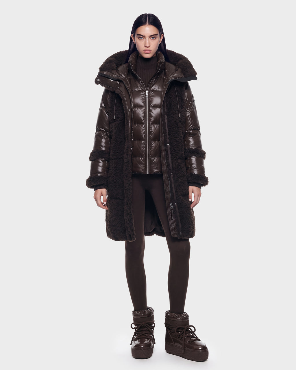 A person stands facing forward against a plain light background, wearing the Rudsak SHELLEY - 8126729 Espresso—a long, dark brown puffer coat with down insulation and fur trim—paired with a matching turtleneck, leggings, and high-top boots for a stylish winter outerwear look.