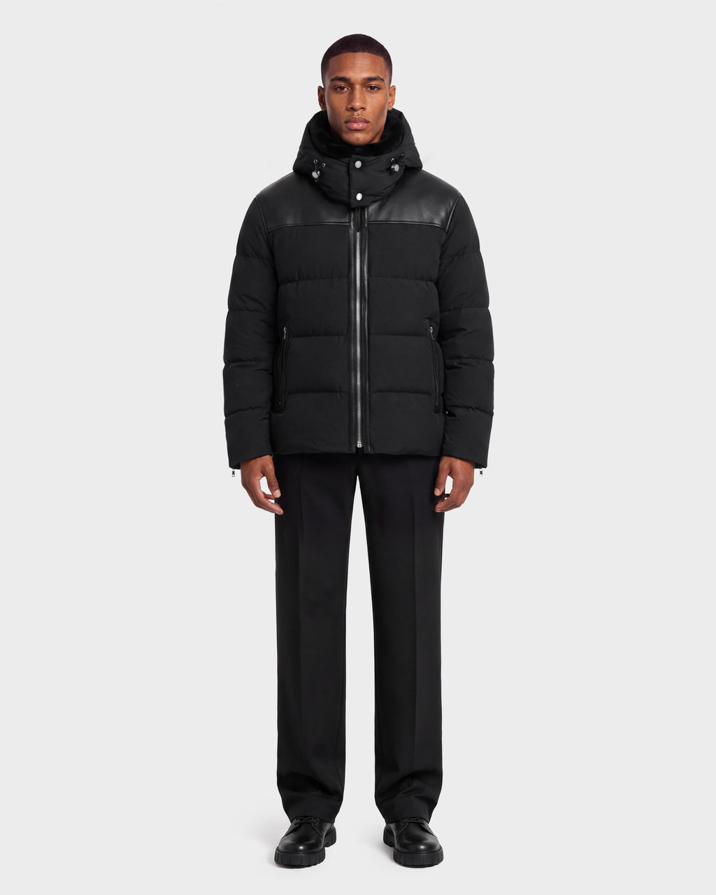 A model wears a black hooded quilted SIMON PX - 6126503 Black puffer jacket by Rudsak with leather shoulder detailing, paired with black trousers and black shoes.