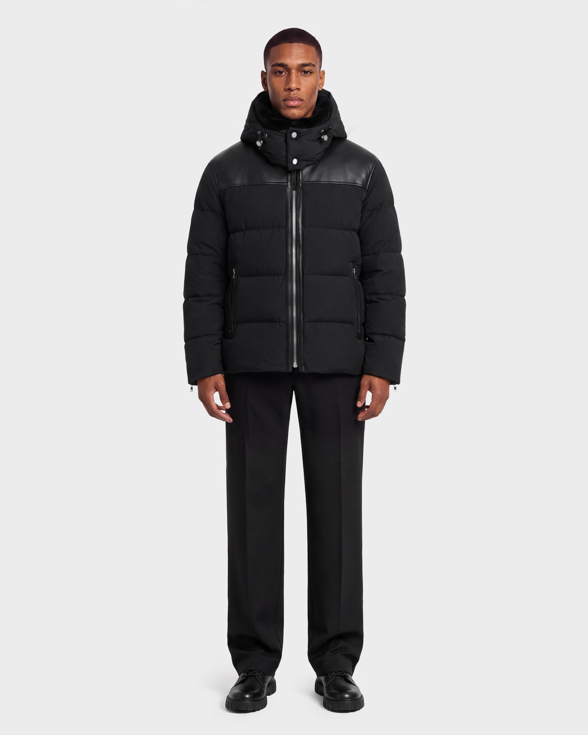 A model wears a black hooded quilted SIMON PX - 6126503 Black puffer jacket by Rudsak with leather shoulder detailing, paired with black trousers and black shoes.