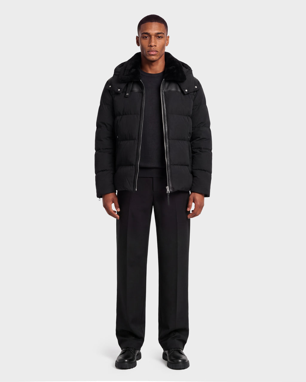A man wearing a black Rudsak SIMON PX - 6126503 Black quilted puffer jacket with a faux fur collar and leather shoulder detailing, paired with a black sweater, black trousers, and black shoes.