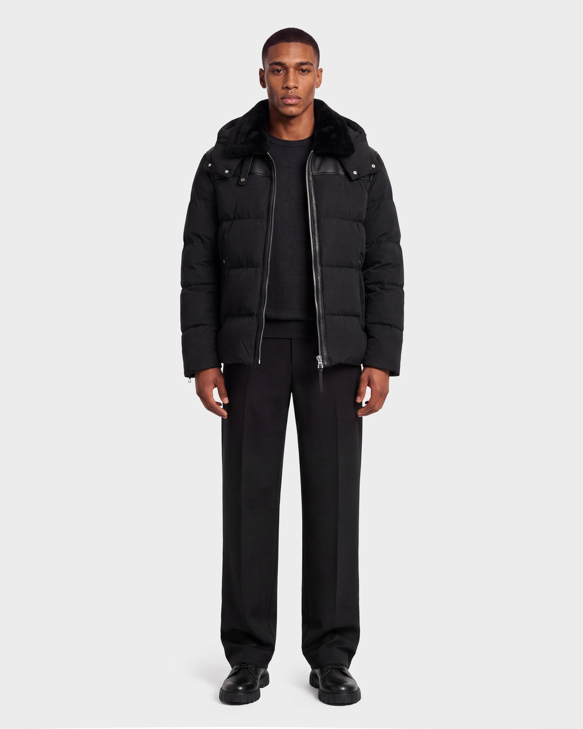 A man wearing a black Rudsak SIMON PX - 6126503 Black quilted puffer jacket with a faux fur collar and leather shoulder detailing, paired with a black sweater, black trousers, and black shoes.