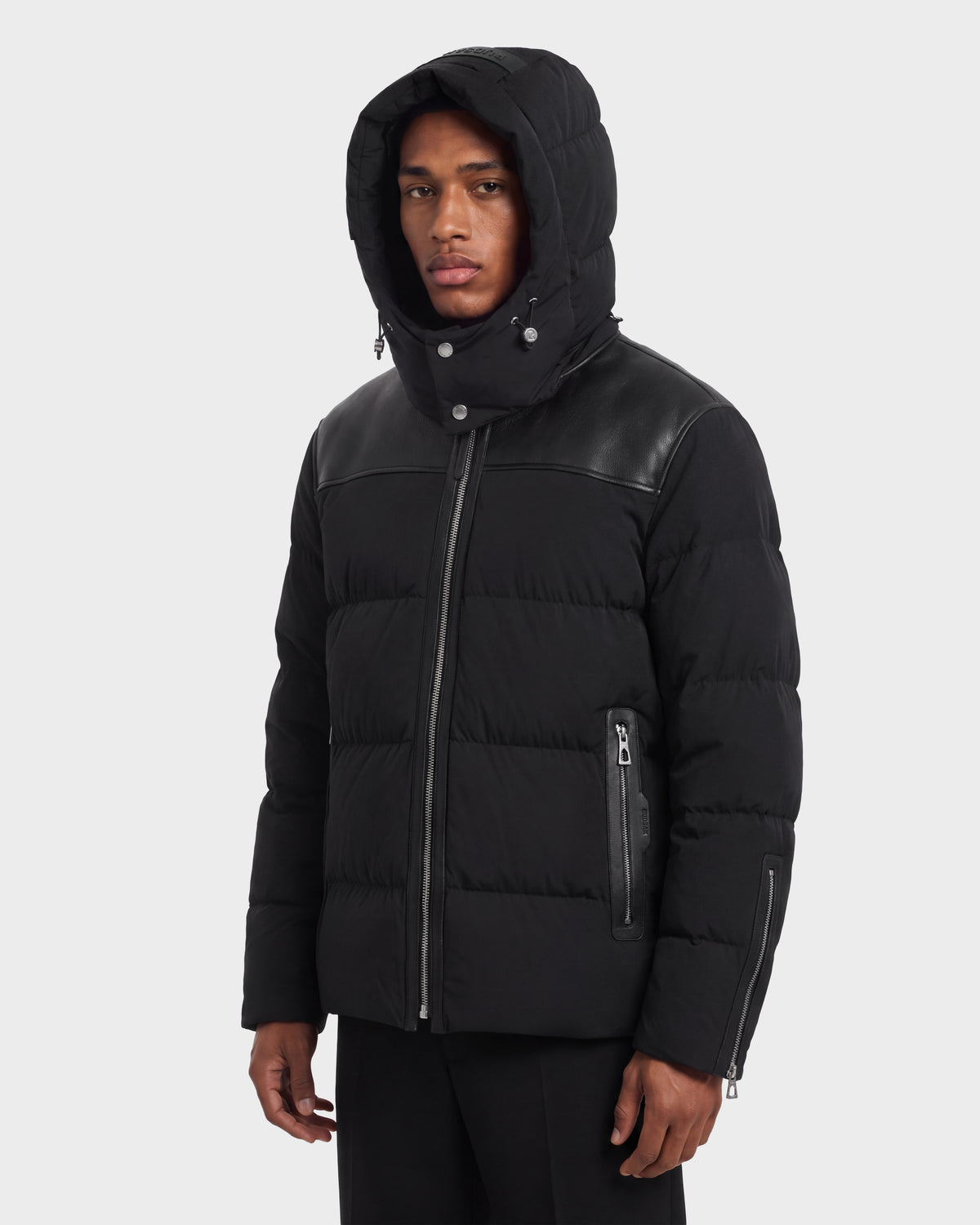 A person wearing a black Rudsak SIMON PX - 6126503 quilted puffer jacket with leather shoulder panels, front zippered pockets, and RDS-certified down insulation.