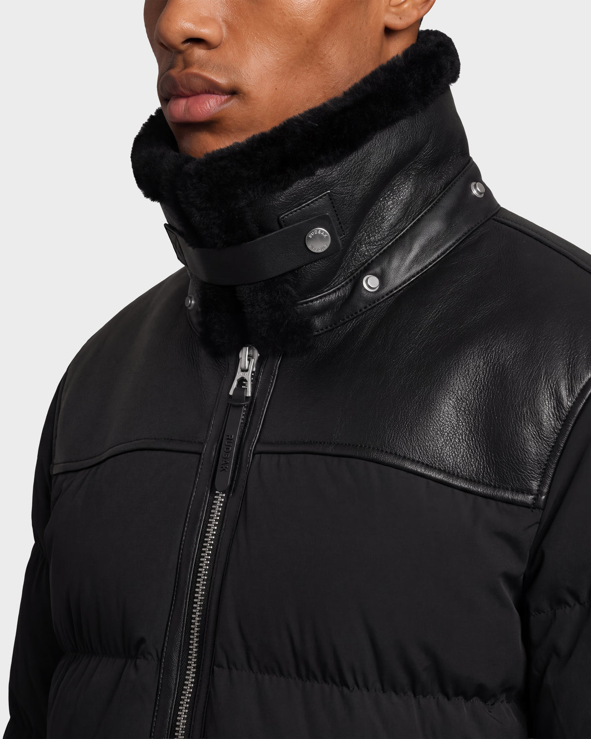 A black quilted Rudsak SIMON PX - 6126503 puffer jacket featuring a leather upper panel and a high collar lined with black fur.