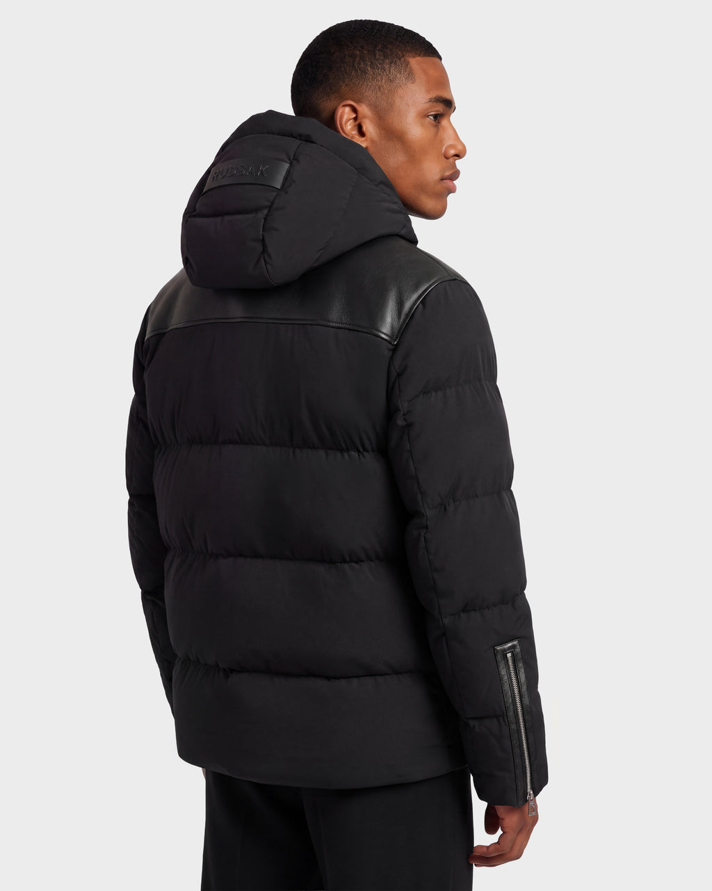 A man wearing a black Rudsak SIMON PX - 6126503 quilted puffer jacket with a hood, leather shoulder panels, and a zippered sleeve detail.