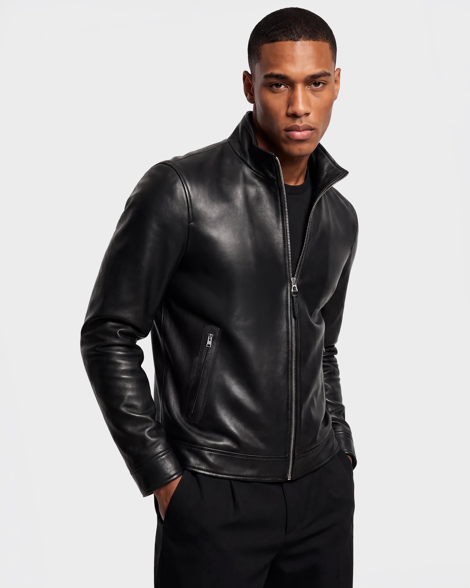 A man wearing the STEVEN - 6124541 Black leather jacket by Rudsak and black pants stands against a plain light background with his hands in his pockets, looking confidently at the camera.