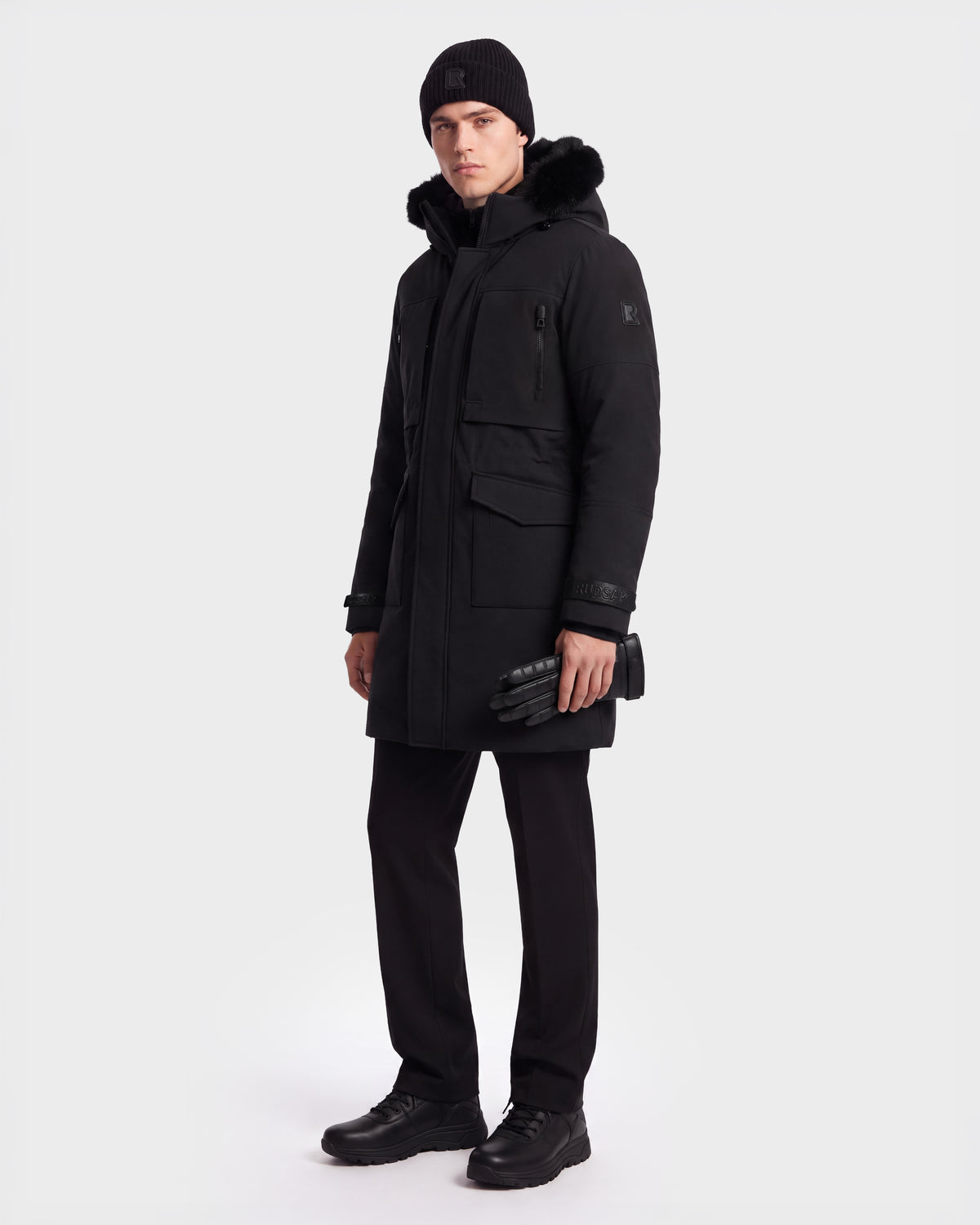 A man stands wearing the Rudsak TROY - 6126504 Black winter parka, paired with black pants, boots, a knit hat, and holding black gloves. The background is plain white.