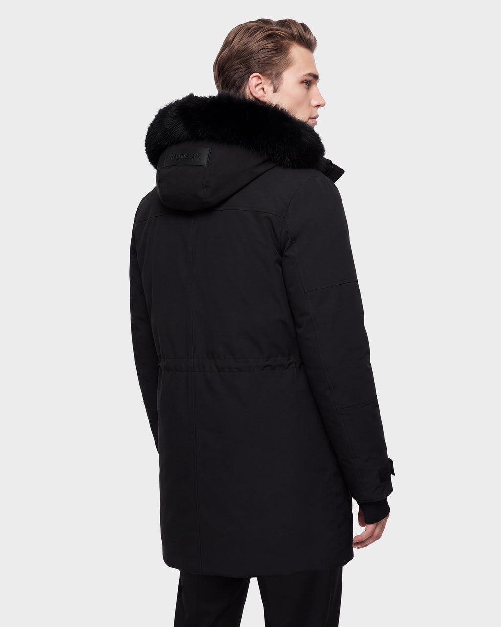 A person is standing and facing away, looking to the side, wearing the Rudsak TROY - 6126504 Black winter coat with a black fur-trimmed hood. The background is plain and light-colored.