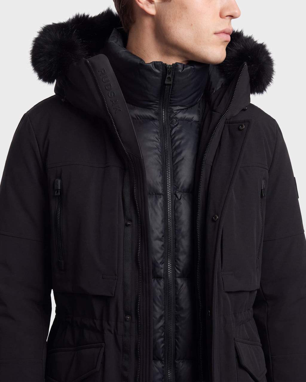 A person wearing the Rudsak TROY - 6126504 Black winter jacket, featuring a fur-lined hood and a high, padded collar, looking to the side against a plain background.