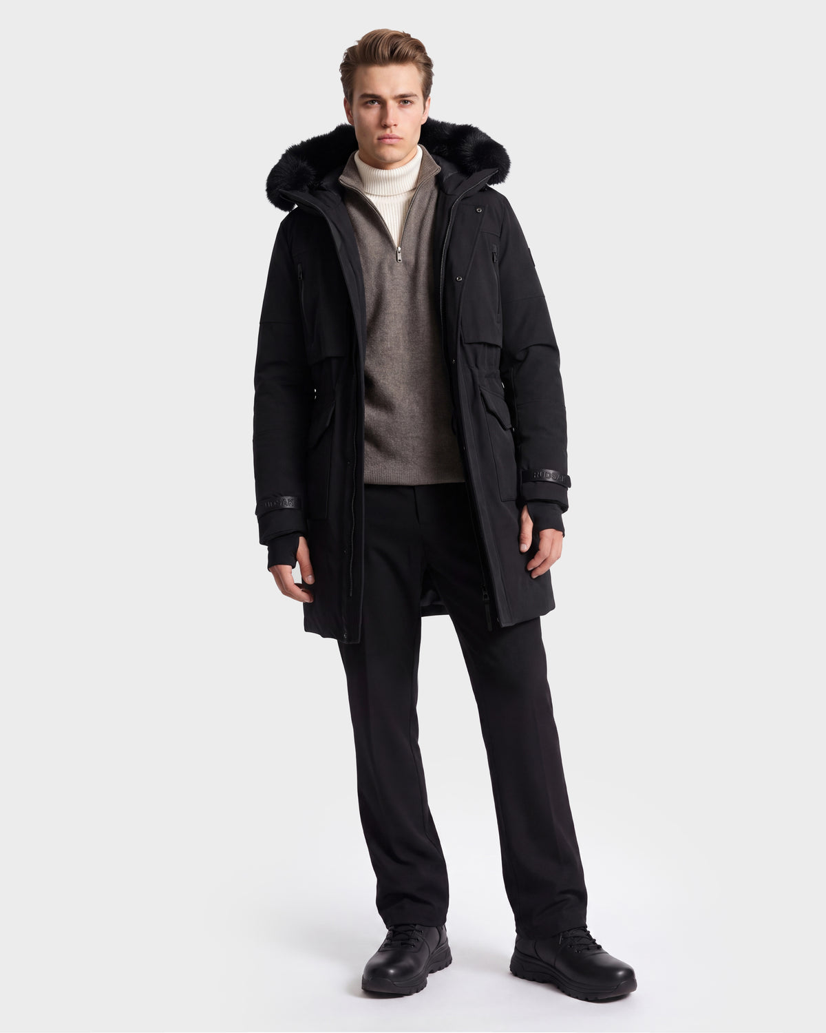 A person stands against a plain white background, wearing the Rudsak TROY - 6126504 Black parka with a fur-lined hood, layered over a gray sweater and cream turtleneck, paired with black pants and black shoes.