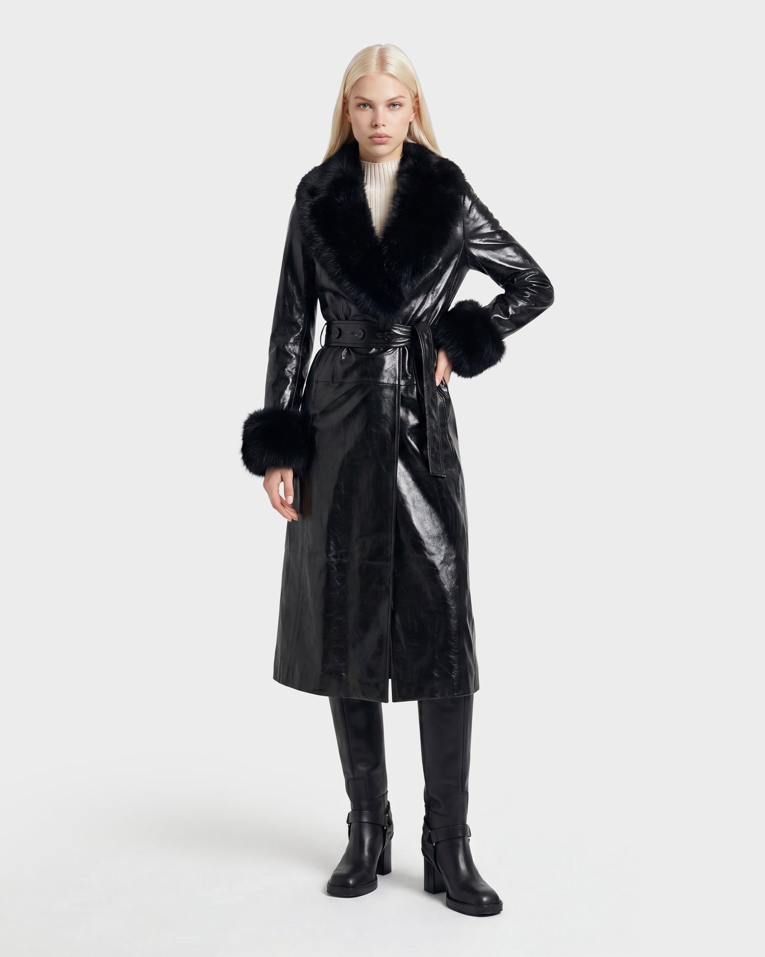 A woman with long blonde hair wears the VIVIANNE - 8126933 Black belted coat by Rudsak, featuring a shiny finish, faux fur collar and cuffs, paired with black knee-high boots and a white turtleneck. She stands against a plain light background. This stylish look is now available for pre-order, with expected delivery beginning August 17, 2026.