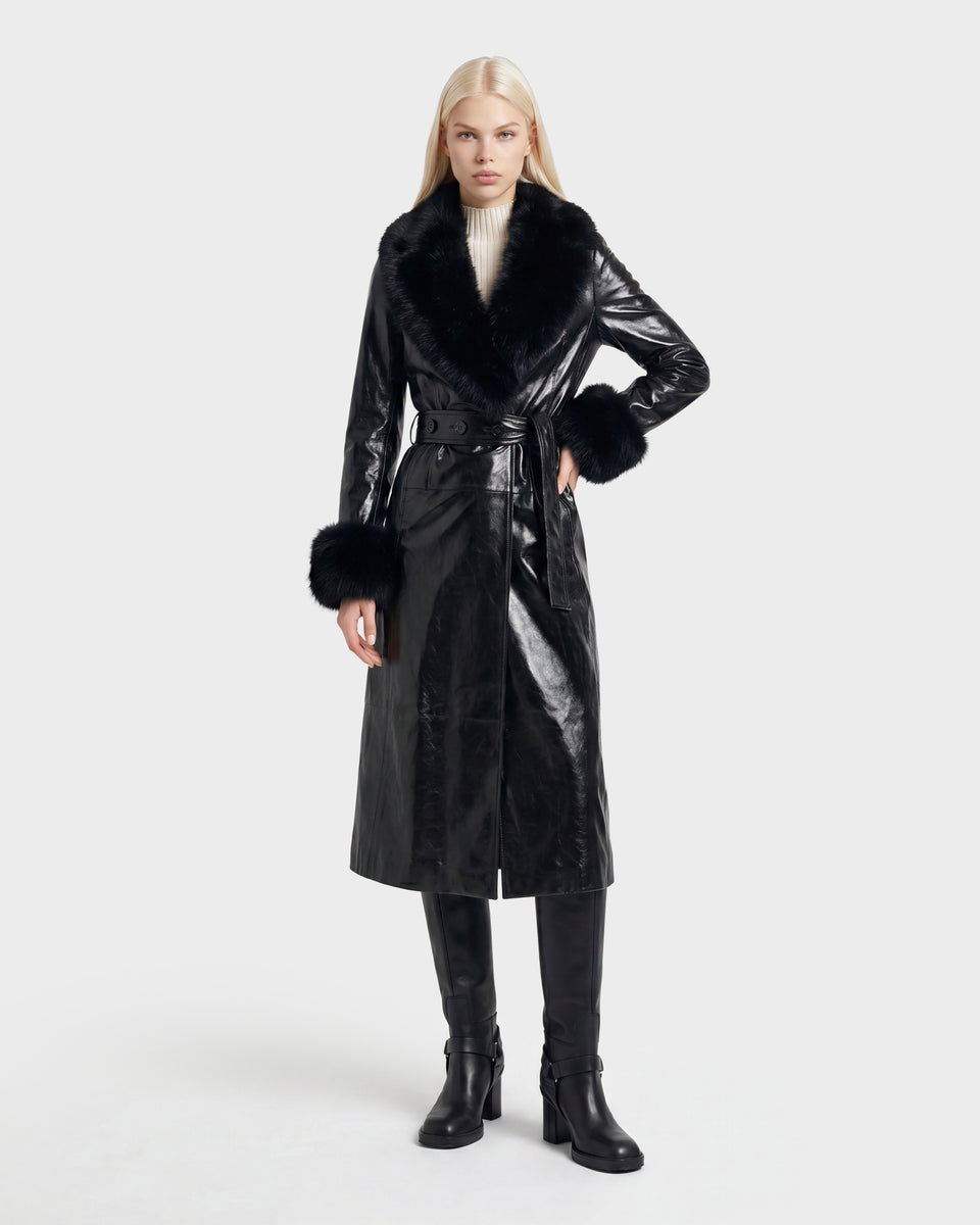 A woman with long blonde hair wears the VIVIANNE - 8126933 Black belted coat by Rudsak, featuring a shiny finish, faux fur collar and cuffs, paired with black knee-high boots and a white turtleneck. She stands against a plain light background. This stylish look is now available for pre-order, with expected delivery beginning August 17, 2026.