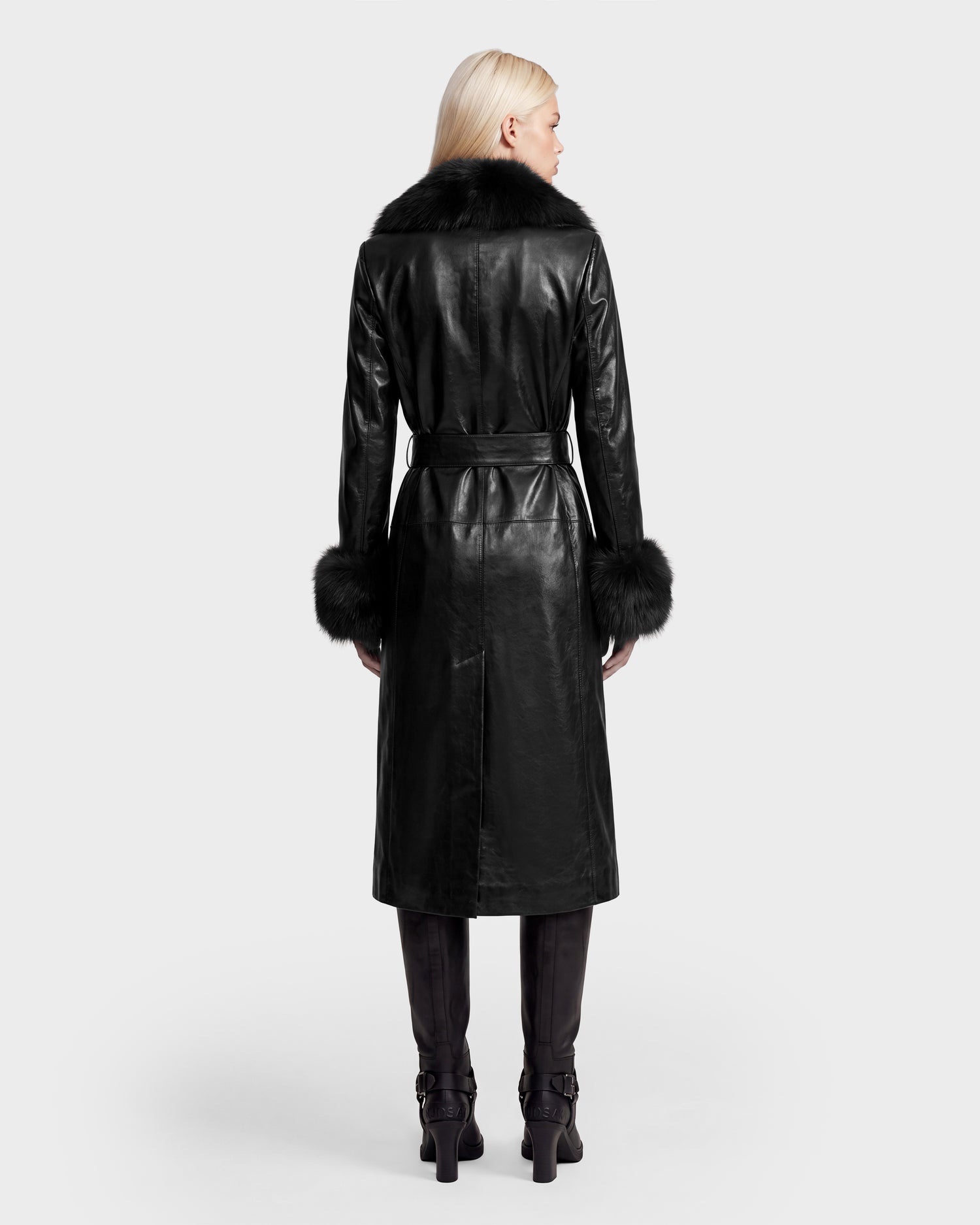 A person with long blonde hair wears the Rudsak VIVIANNE - 8126933 Black leather trench coat, featuring fur trim on the collar and cuffs, paired with black pants and black boots. They are standing against a white background, viewed from the back. This striking look is available for pre-order now, with expected delivery by August 17, 2026.