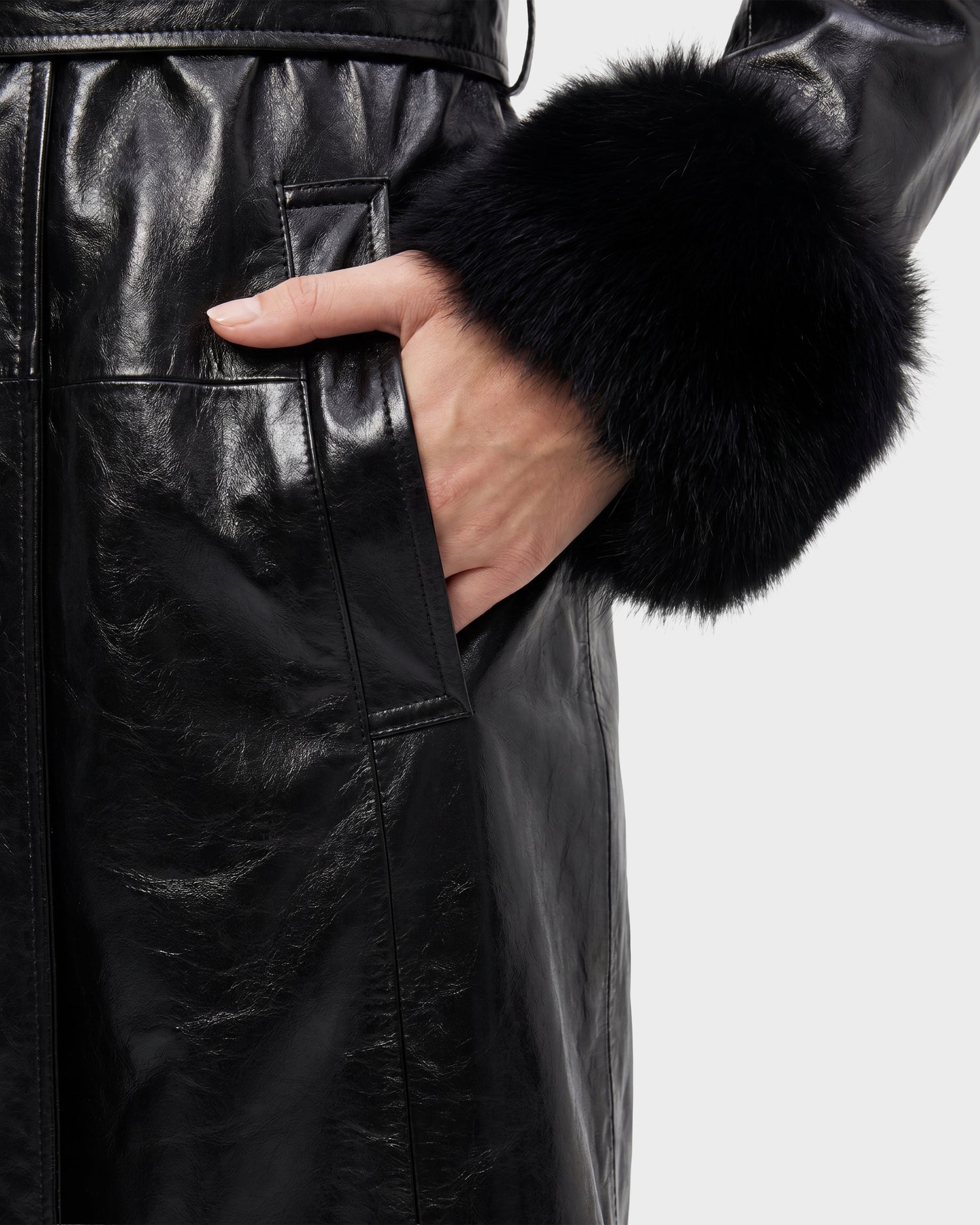 A close-up of a person’s hand in the pocket of the VIVIANNE - 8126933 Black coat by Rudsak, featuring shiny black leather and black fur trim on the sleeve, now available for pre-order with expected delivery by August 17, 2026.