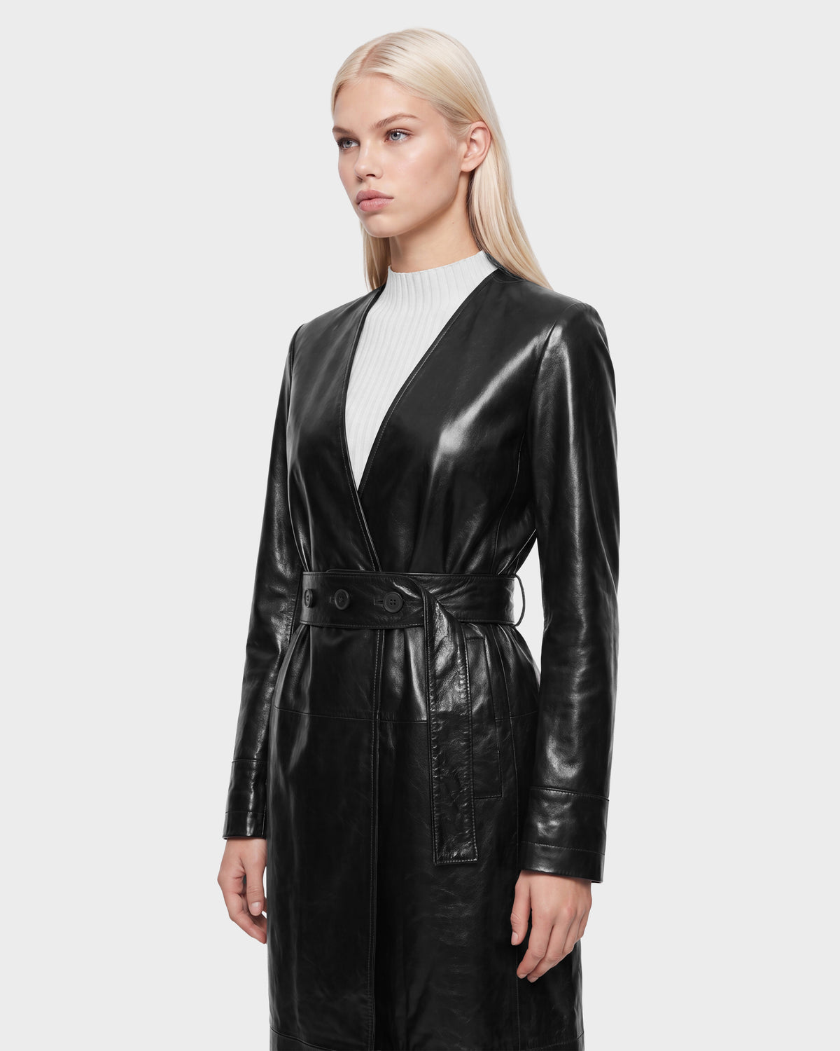 A person with long blonde hair wears the Rudsak VIVIANNE - 8126933 Black belted leather coat over a white ribbed top, standing against a plain light gray background—an effortlessly chic look now available for pre-order, with expected delivery by August 17, 2026.