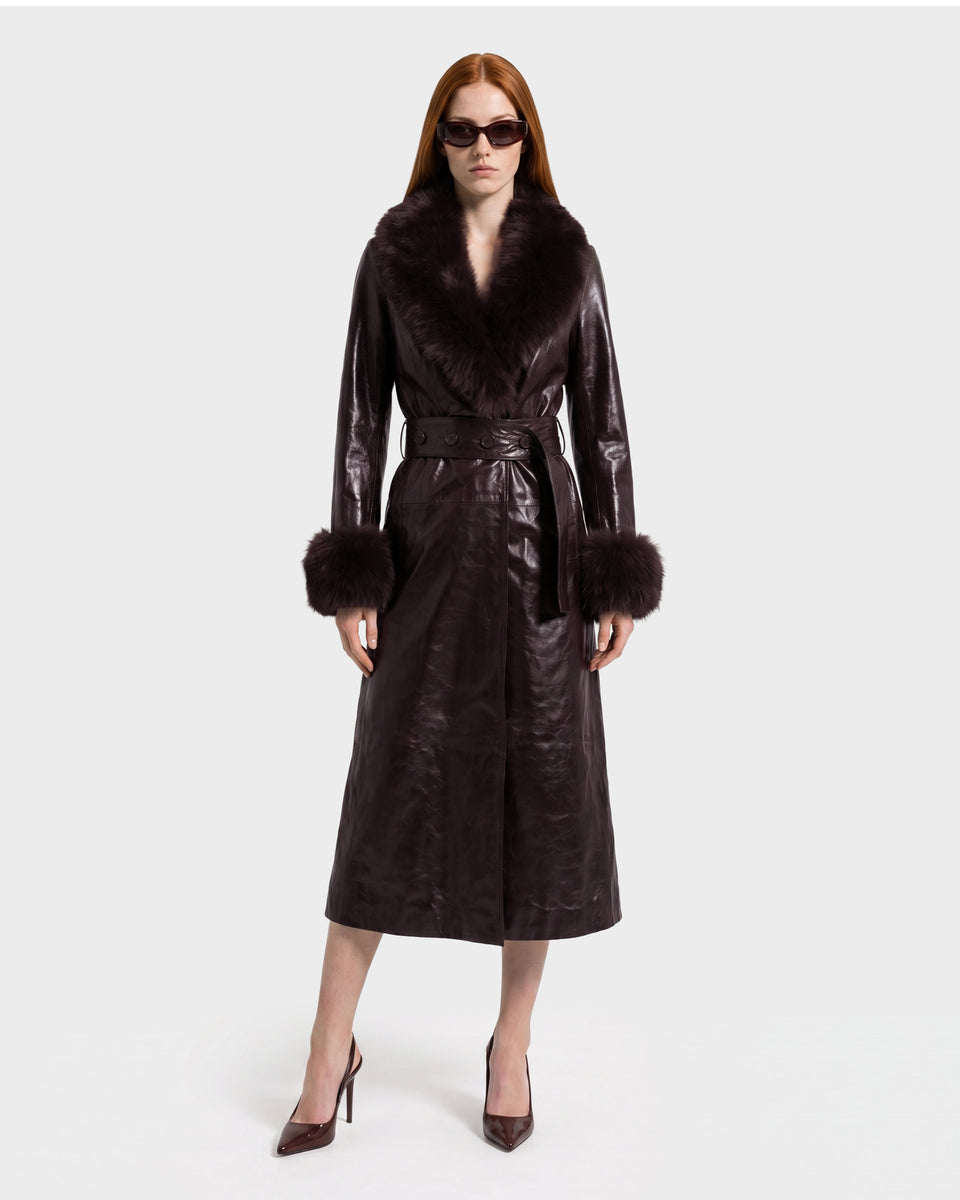 A woman wearing the Rudsak VIVIANNE - 8126933 Espresso, a dark brown belted leather coat with fur trim on the collar and cuffs, stands against a plain light background in matching heels.