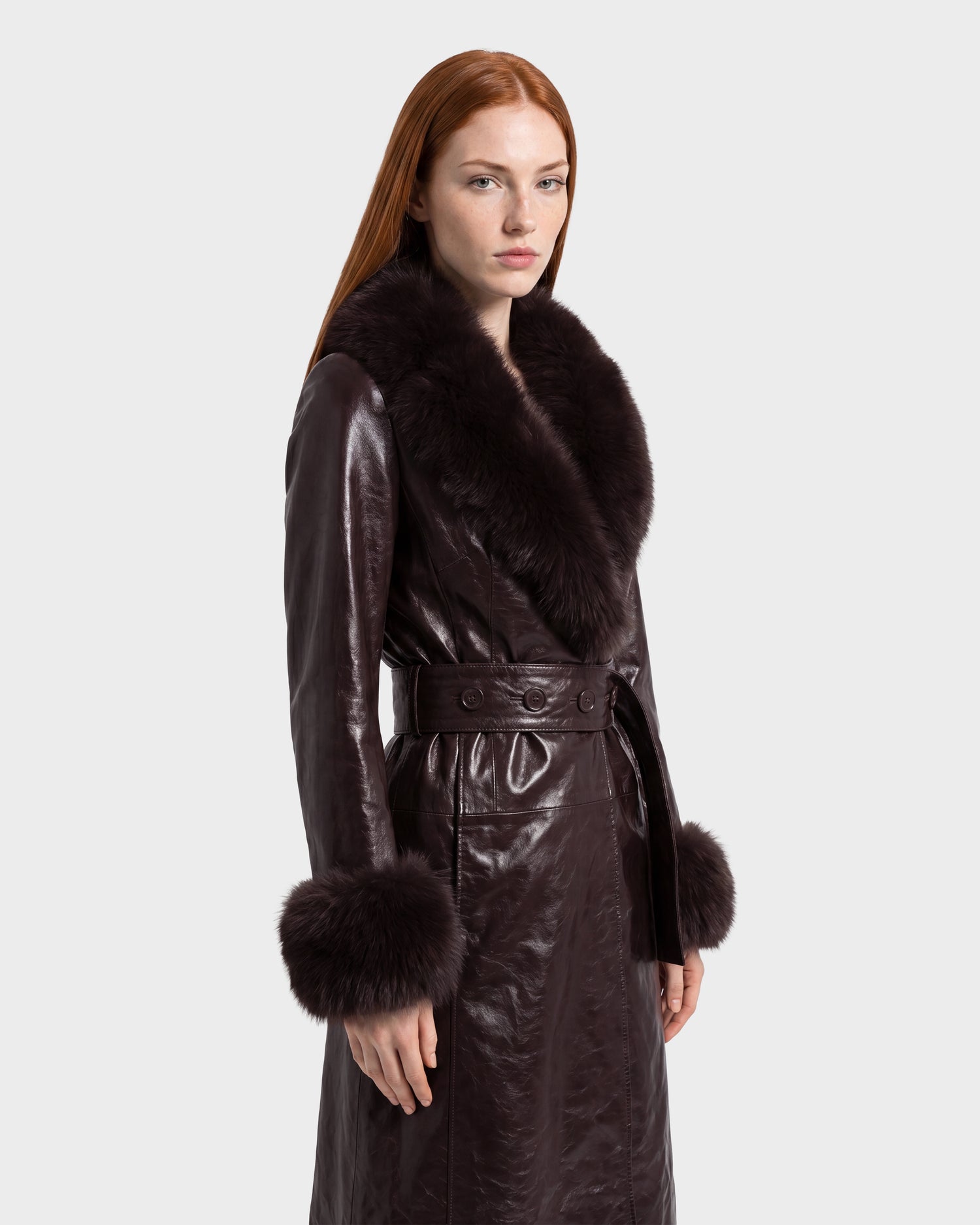 A woman with long red hair wears the Rudsak VIVIANNE - 8126933 Espresso dark brown leather coat, featuring large, fluffy fur collar and cuffs, as she stands against a plain light background.
