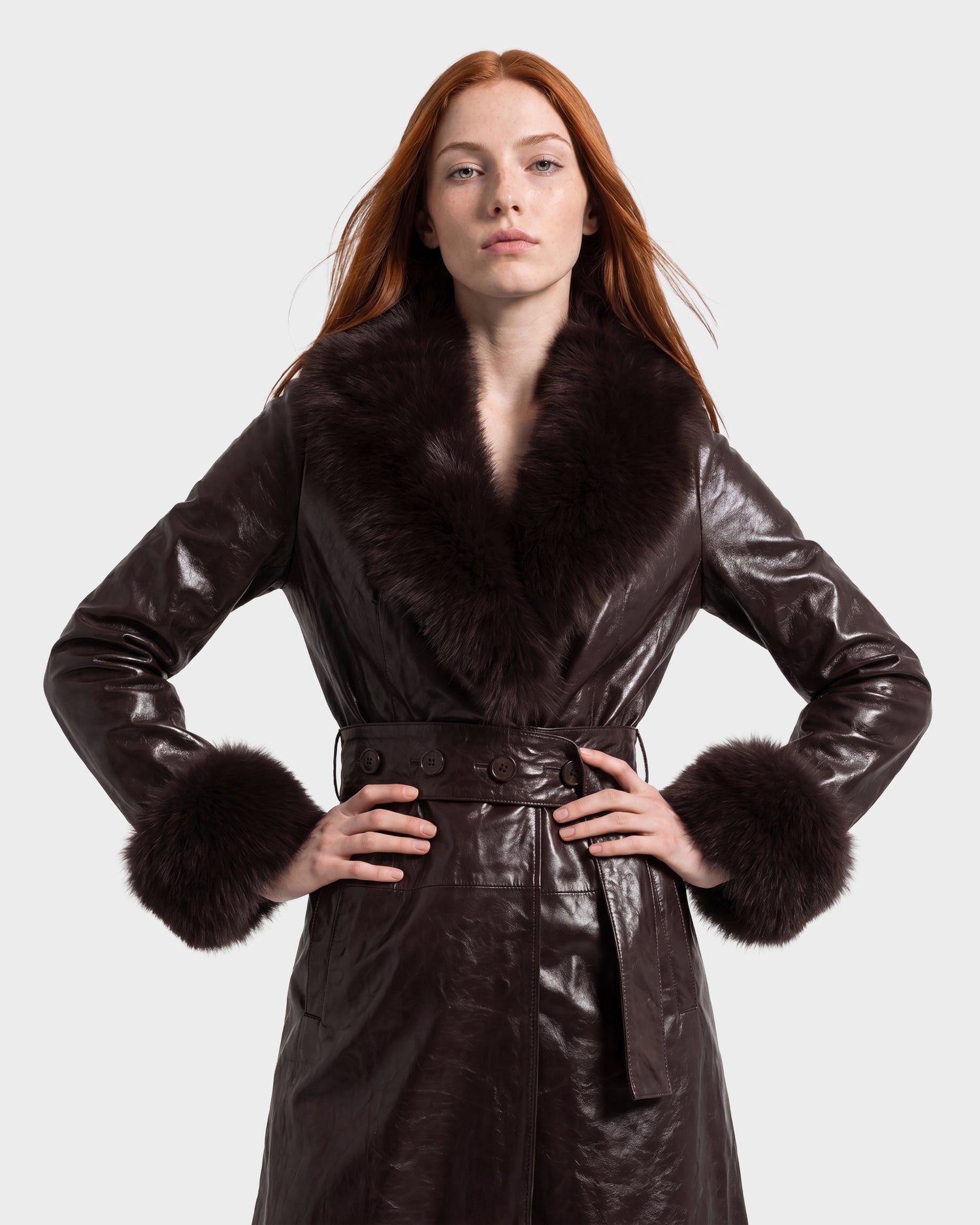 A person with long red hair stands with hands on hips against a plain light background, wearing the Rudsak VIVIANNE - 8126933 Espresso dark brown leather coat with fur collar and cuffs.