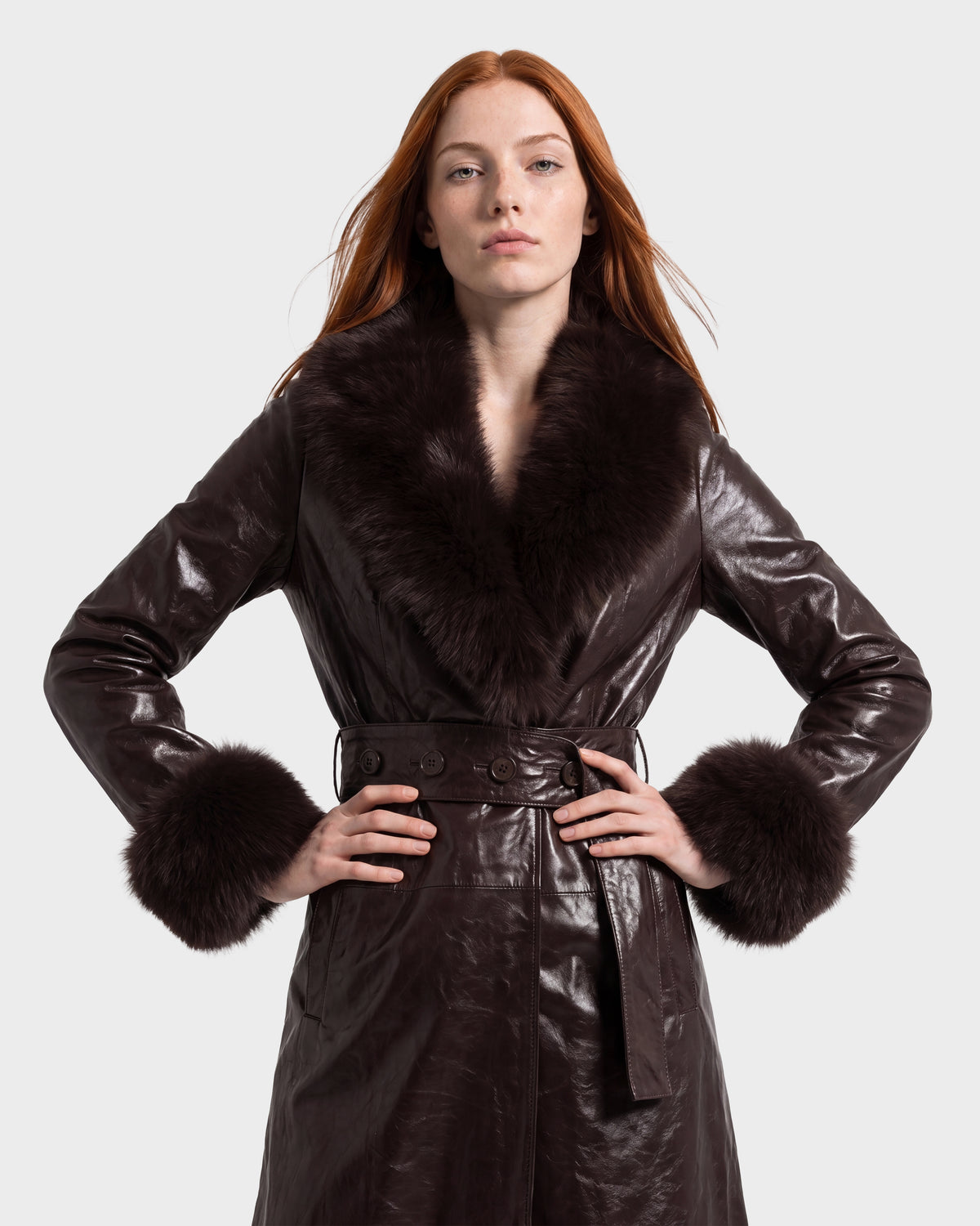 A person with long red hair stands with hands on hips against a plain light background, wearing the Rudsak VIVIANNE - 8126933 Espresso dark brown leather coat with fur collar and cuffs.