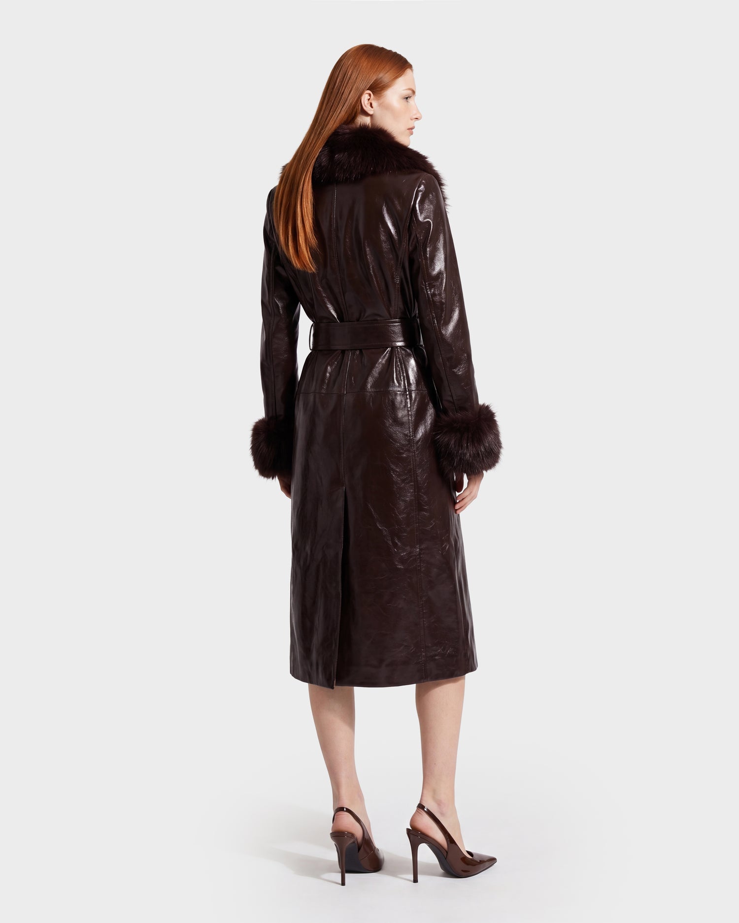 A woman with long red hair wears the Rudsak VIVIANNE - 8126933 Espresso dark brown leather coat, featuring a fur collar and cuffs, paired with high heels. She is standing and facing away from the camera against a plain, light background.