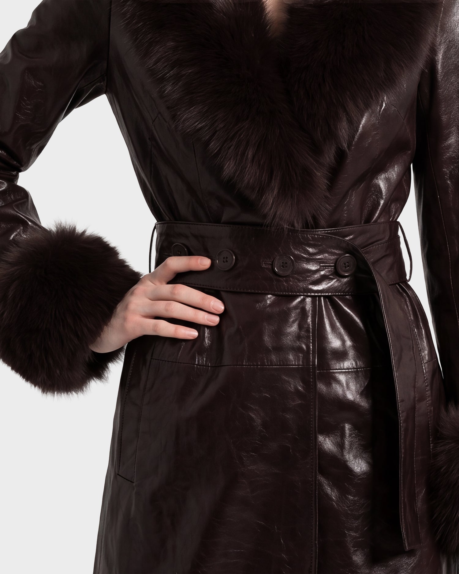 A person wearing the Rudsak VIVIANNE - 8126933 Espresso, a dark brown leather coat with a fur collar and cuffs, rests one hand on their waist, showcasing the button and belt details.