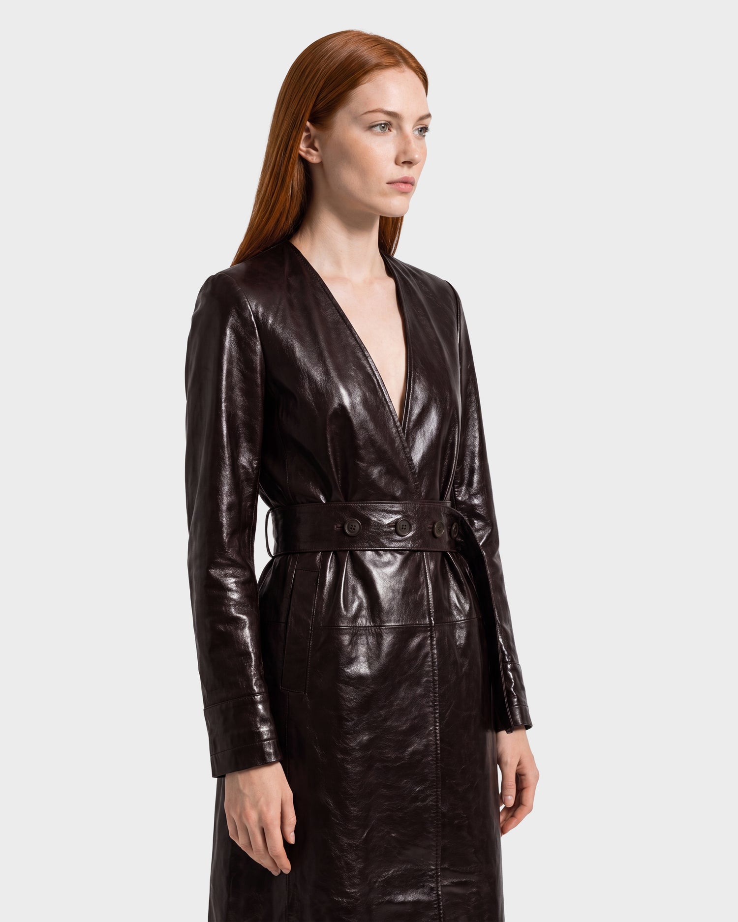 A woman with long red hair wears the Rudsak VIVIANNE - 8126933 Espresso belted leather coat, standing against a plain light gray background and looking slightly to the side with a neutral expression.