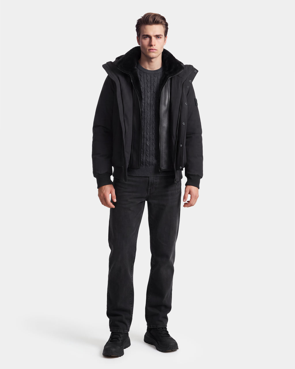 A man stands facing forward wearing the Rudsak WILL - 6126561 Black jacket with a fur collar, styled with a dark cable-knit sweater, black jeans, and black shoes against a plain white background.