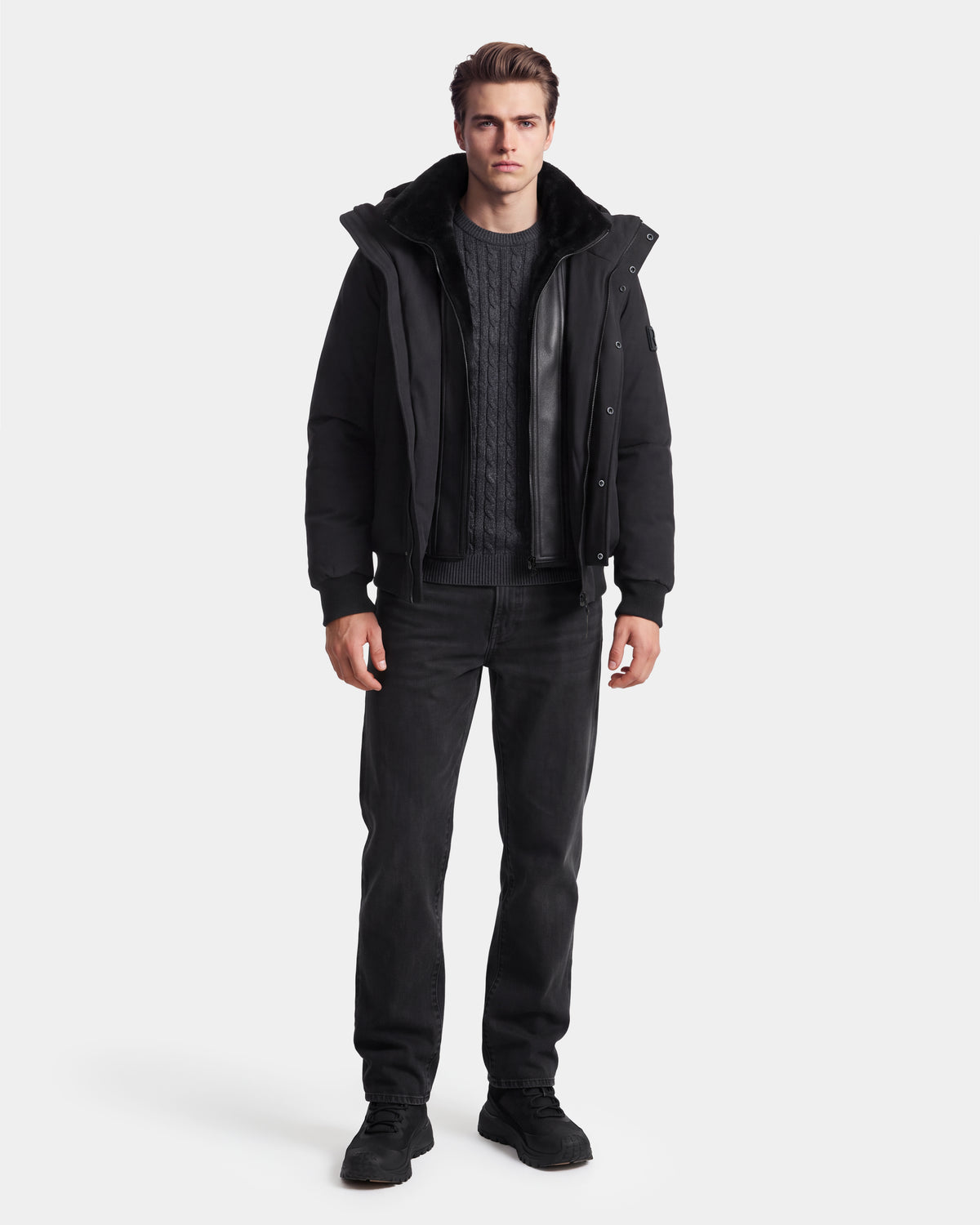 A man stands facing forward wearing the Rudsak WILL - 6126561 Black jacket with a fur collar, styled with a dark cable-knit sweater, black jeans, and black shoes against a plain white background.