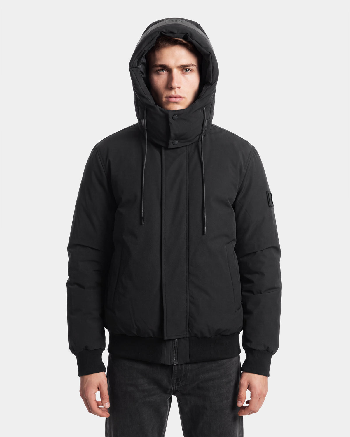A person wearing the Rudsak WILL - 6126561 Black hooded winter jacket with a high collar stands facing forward with a neutral expression against a plain white background.