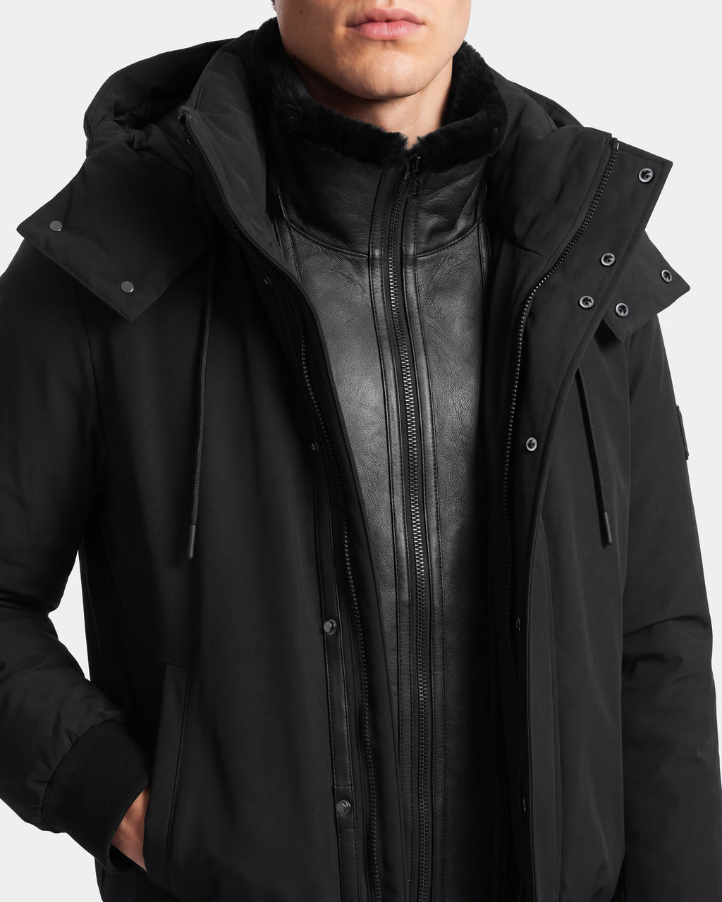 A person wearing the Rudsak WILL - 6126561 Black hooded jacket layered over a black leather zip-up jacket, shown from the shoulders to the mouth against a plain background.