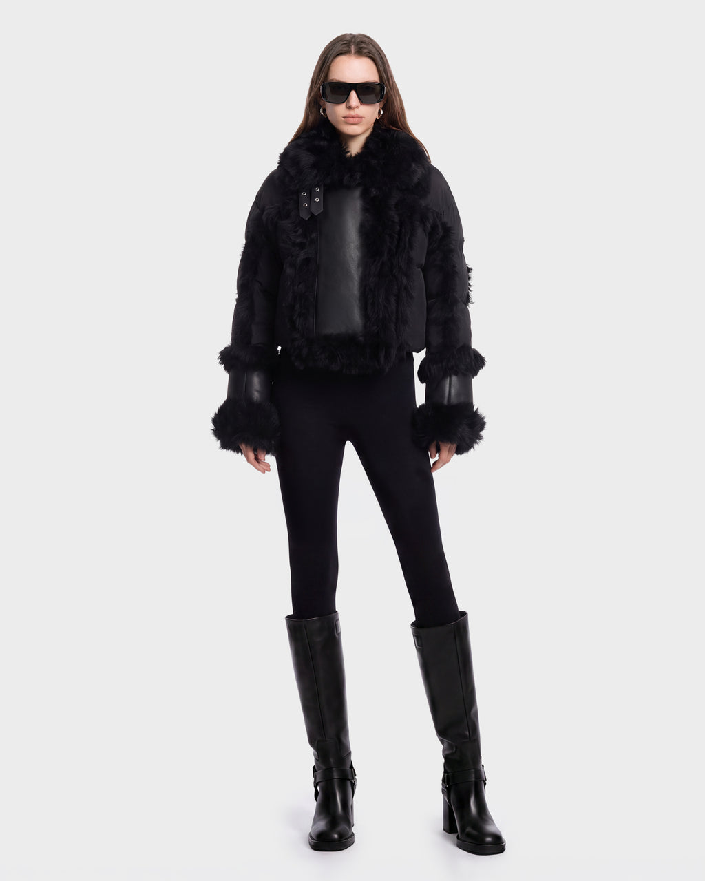 A person stands facing forward against a plain white background wearing the ZELAIA - 8125577 Black cropped puffer with faux-fur detailing by Rudsak, paired with black sunglasses, black leggings, and knee-high black boots.