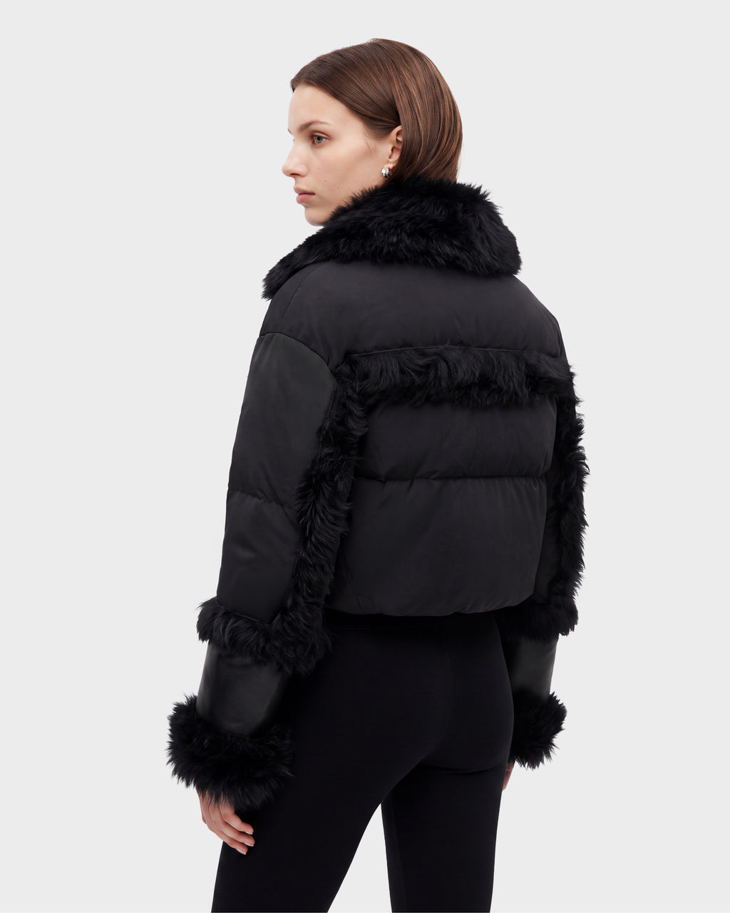 A woman stands facing away, looking over her shoulder against a plain light background, wearing the Rudsak ZELAIA - 8125577 Black cropped puffer jacket with faux-fur detailing and black pants.