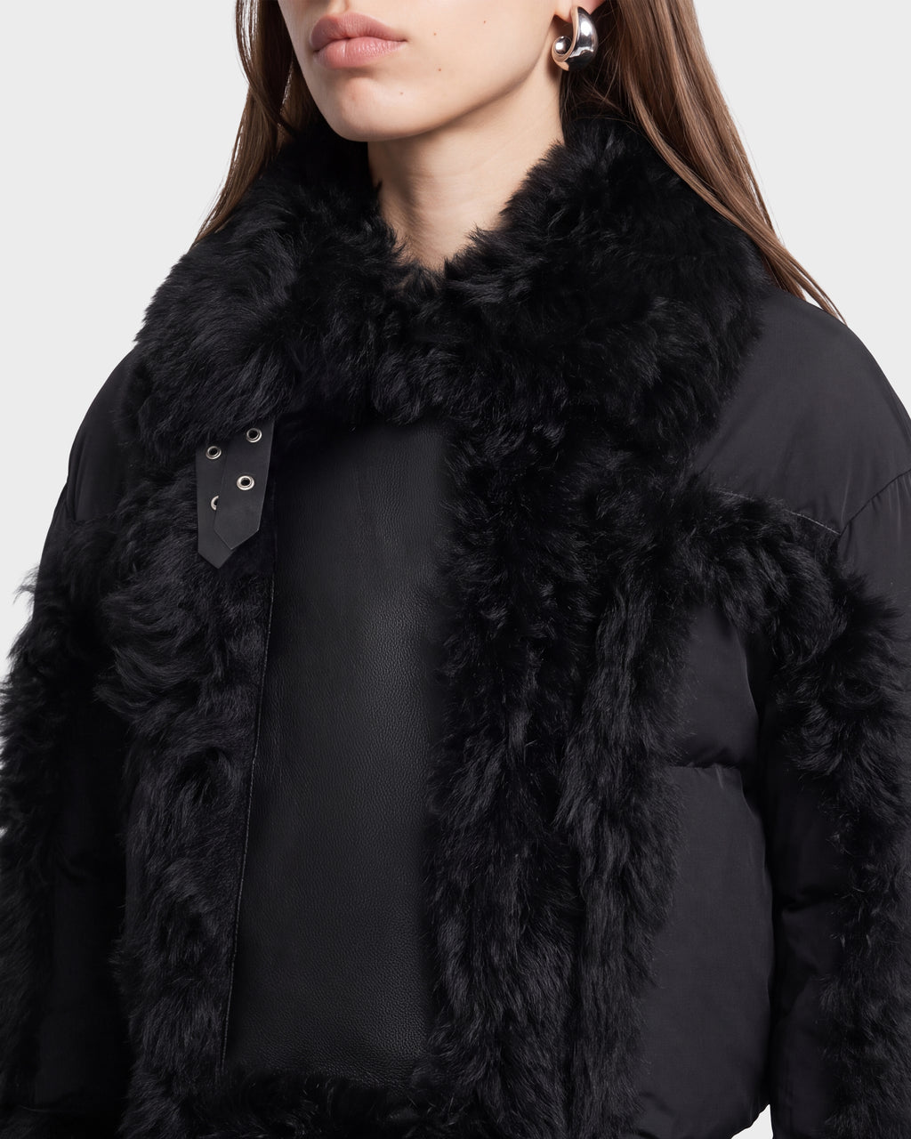 A woman wearing the Rudsak ZELAIA - 8125577 Black coat, featuring large, fluffy faux-fur detailing on the collar and trim, along with silver earrings and straight brown hair, shown from the shoulders up against a plain background.