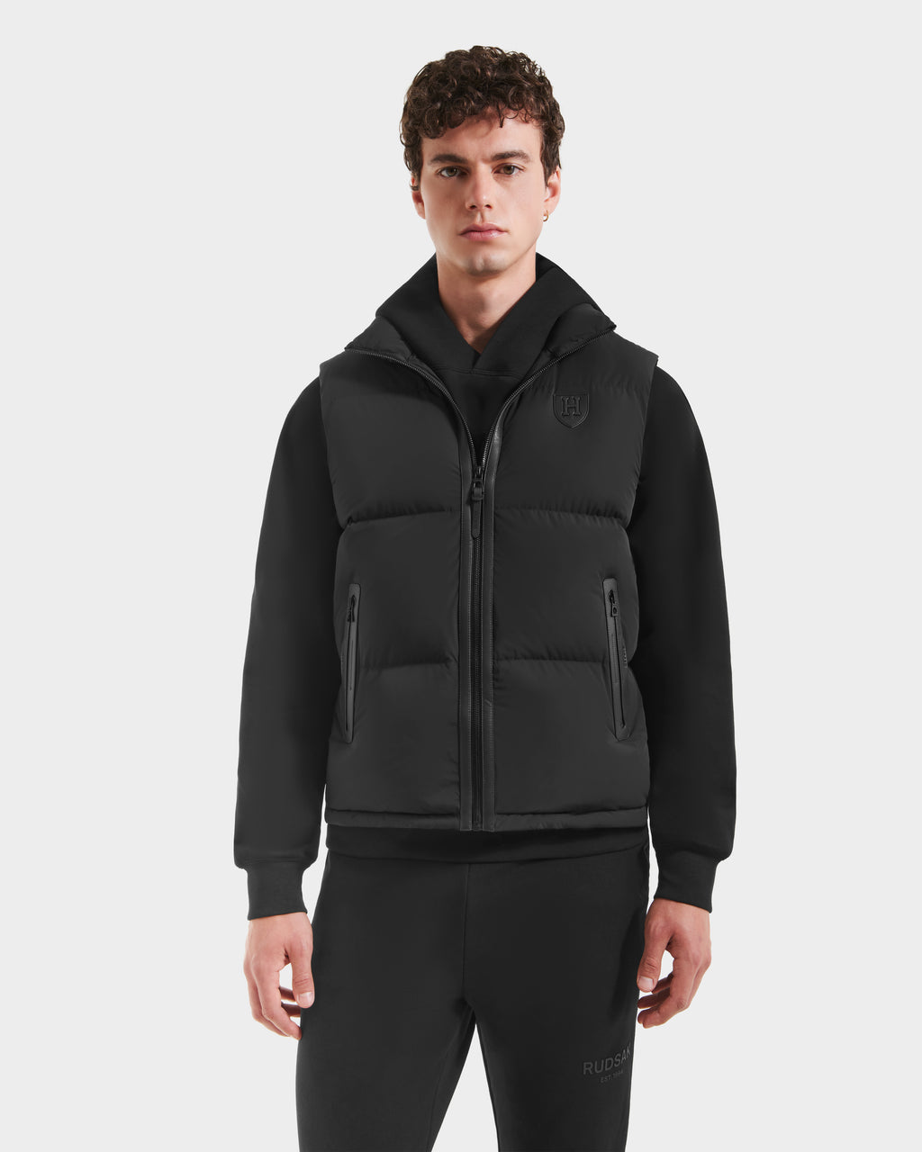 A young man with short curly hair wears the Rudsak FREDDY HV - H125510 Black quilted down puffer vest over a black hoodie and matching pants, standing against a plain white background.