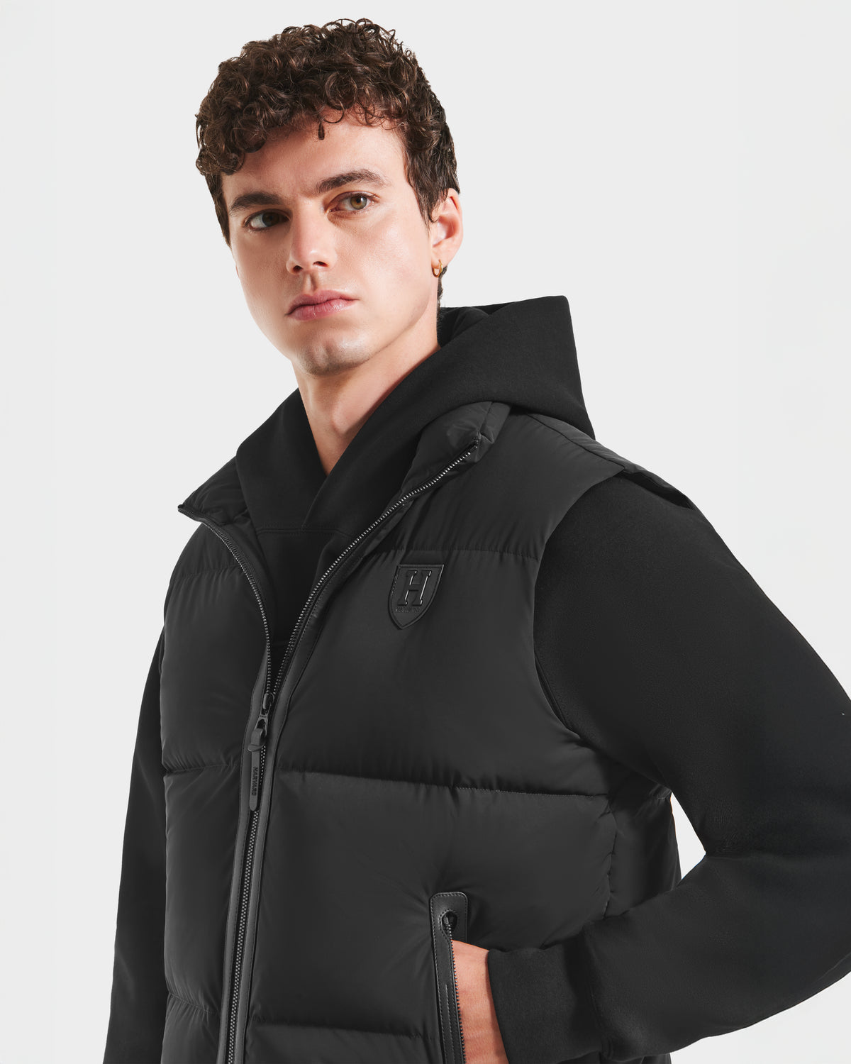 A young man with curly hair wears the Rudsak FREDDY HV - H125510 Black puffer vest, featuring a chest logo, over a black hooded sweatshirt, standing against a plain white background with his hand in his pocket.