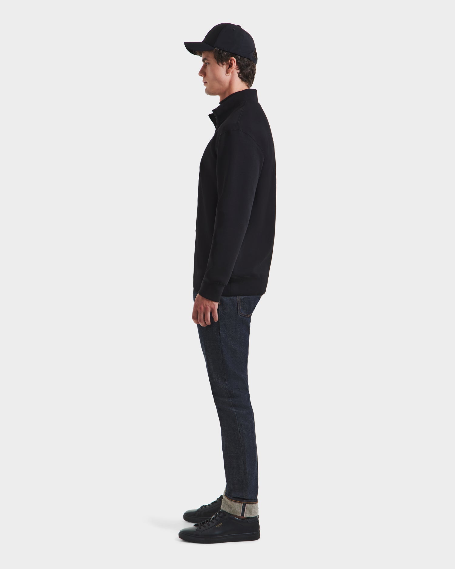 A person stands in profile against a white background, wearing a black baseball cap, the Rudsak FREMONT HV - H525501 Black zip-up jacket made from premium cotton blend, dark cuffed jeans, and black sneakers.
