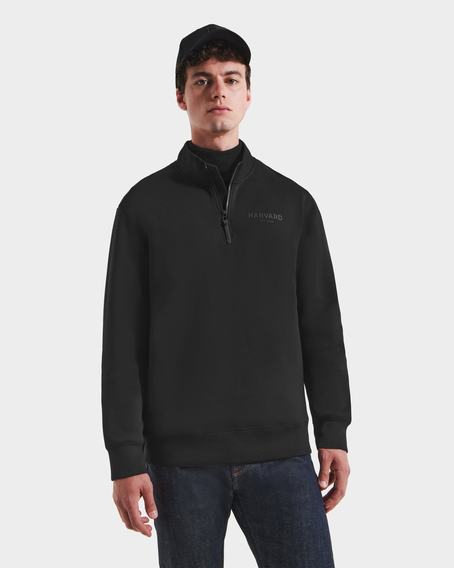 A young man wearing a Rudsak FREMONT HV - H525501 Black quarter-zip sweatshirt, dark jeans, and a black cap stands facing forward against a plain white background.