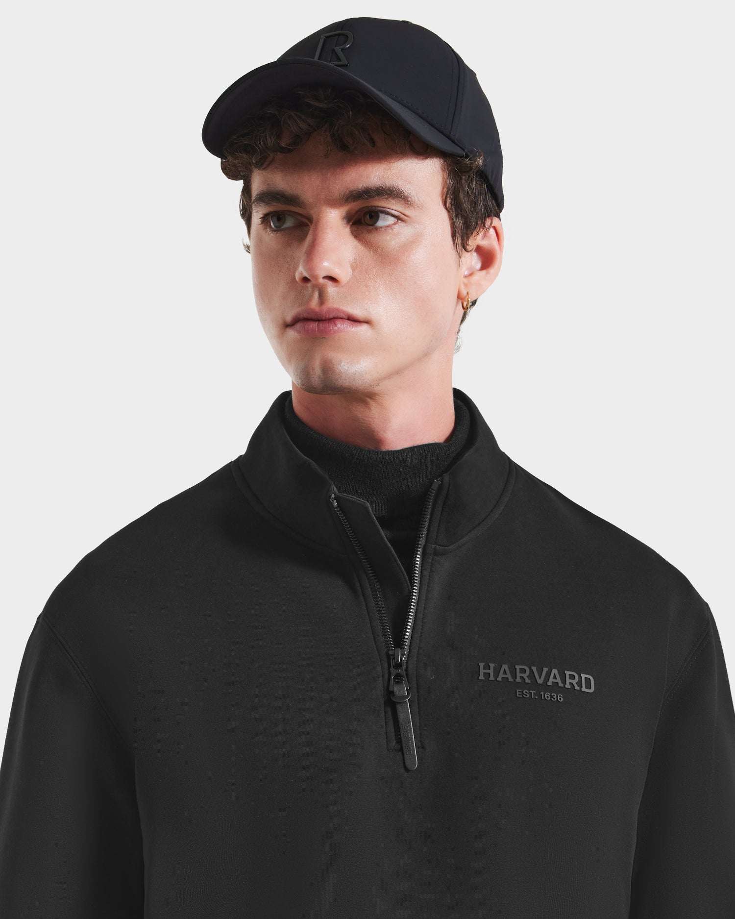 A young man in the Rudsak FREMONT HV - H525501 Black zip-up sweatshirt, featuring HARVARD EST. 1636 and crafted from a premium cotton blend, paired with a black cap, looks slightly to the side against a white background.