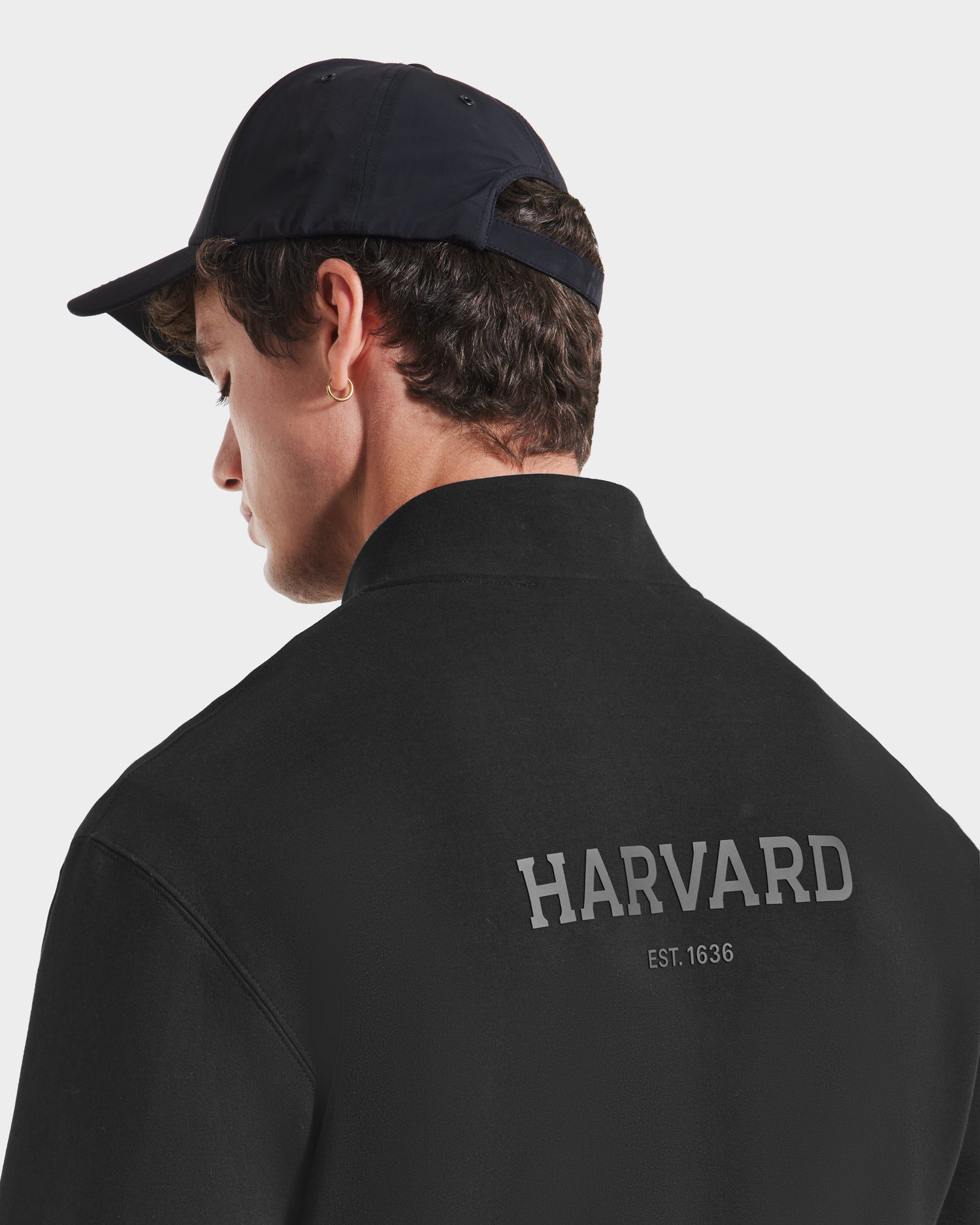 A person wearing a black cap and a Rudsak FREMONT HV - H525501 Black premium cotton shirt with HARVARD EST. 1636 in gray on the back, viewed from behind against a plain background.