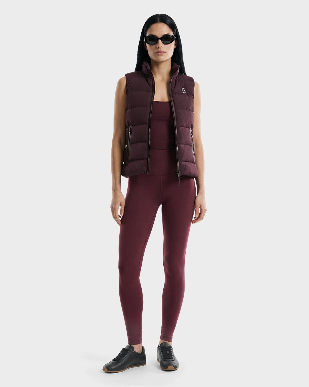 A woman faces forward, wearing Rudsak’s GABRIELA - 8125014 Bordeaux women’s down puffer vest over a matching maroon tank top and leggings with black sunglasses and sneakers. She stands against a plain gray background.