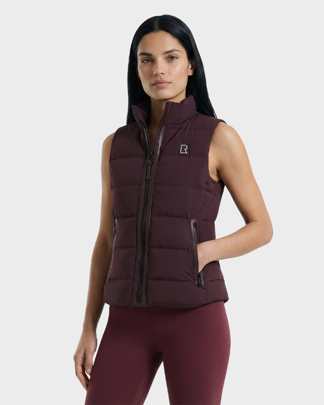 A woman with long dark hair wears the Rudsak GABRIELA - 8125014 Bordeaux women’s down puffer vest with matching leggings, standing against a plain light gray background.