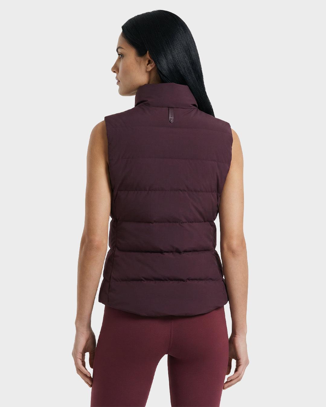A woman with long dark hair, seen from the back, wears the Rudsak GABRIELA - 8125014 Bordeaux sleeveless down puffer vest and matching leggings against a plain light gray background.