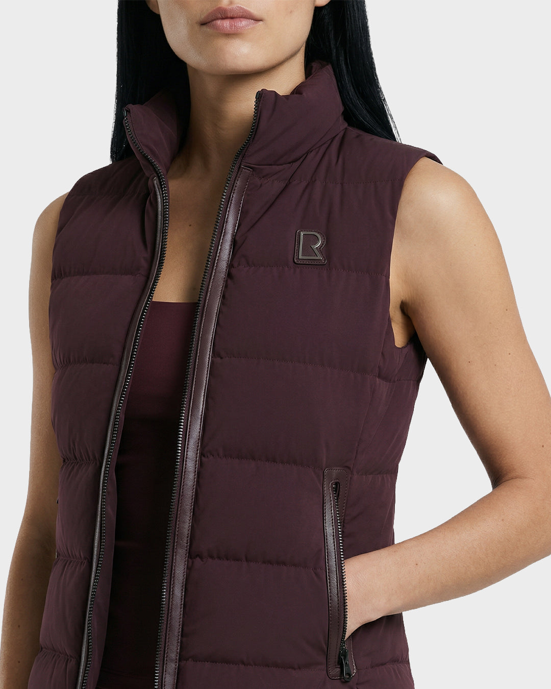 A woman wears the Rudsak GABRIELA - 8125014 Bordeaux sleeveless down puffer vest with a stand collar, zip front, logo patch on the left chest, and her right hand in the zippered pocket.