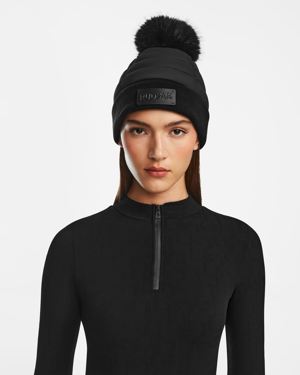 A woman wears the Rudsak GALA - 8424514 Black zip-up top and a black winter hat with a faux fur pom pom and Rudsak logo, standing against a plain white background.